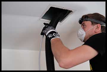 Duct cleaning Monmouth County being done by man wearing work gloves, white respiratory mask, head lamp and black t-shirt; has long black hose inserted into a duct in the ceiling